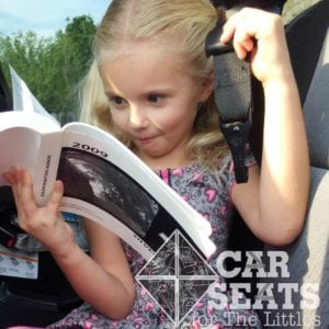 Preschool aged child holding car seat tether in one hand while reading vehicle manual and making a goofy face.